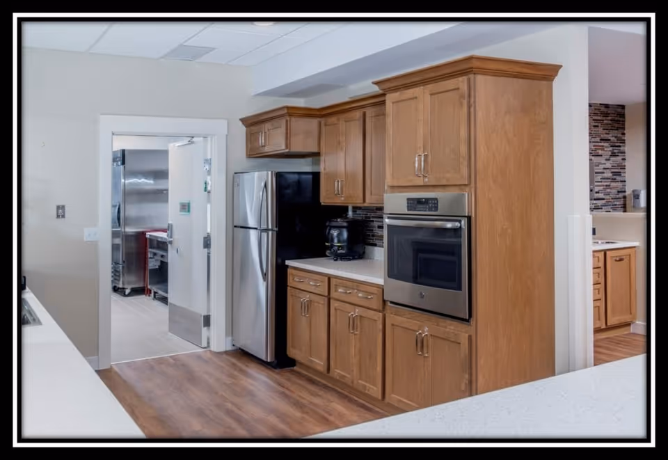 Interior view of a kitchen area with wooden cabinets, a stainless steel refrigerator, and a built-in oven. The floor is wooden, and there is a doorway leading to another room with stainless steel appliances and tiled flooring.