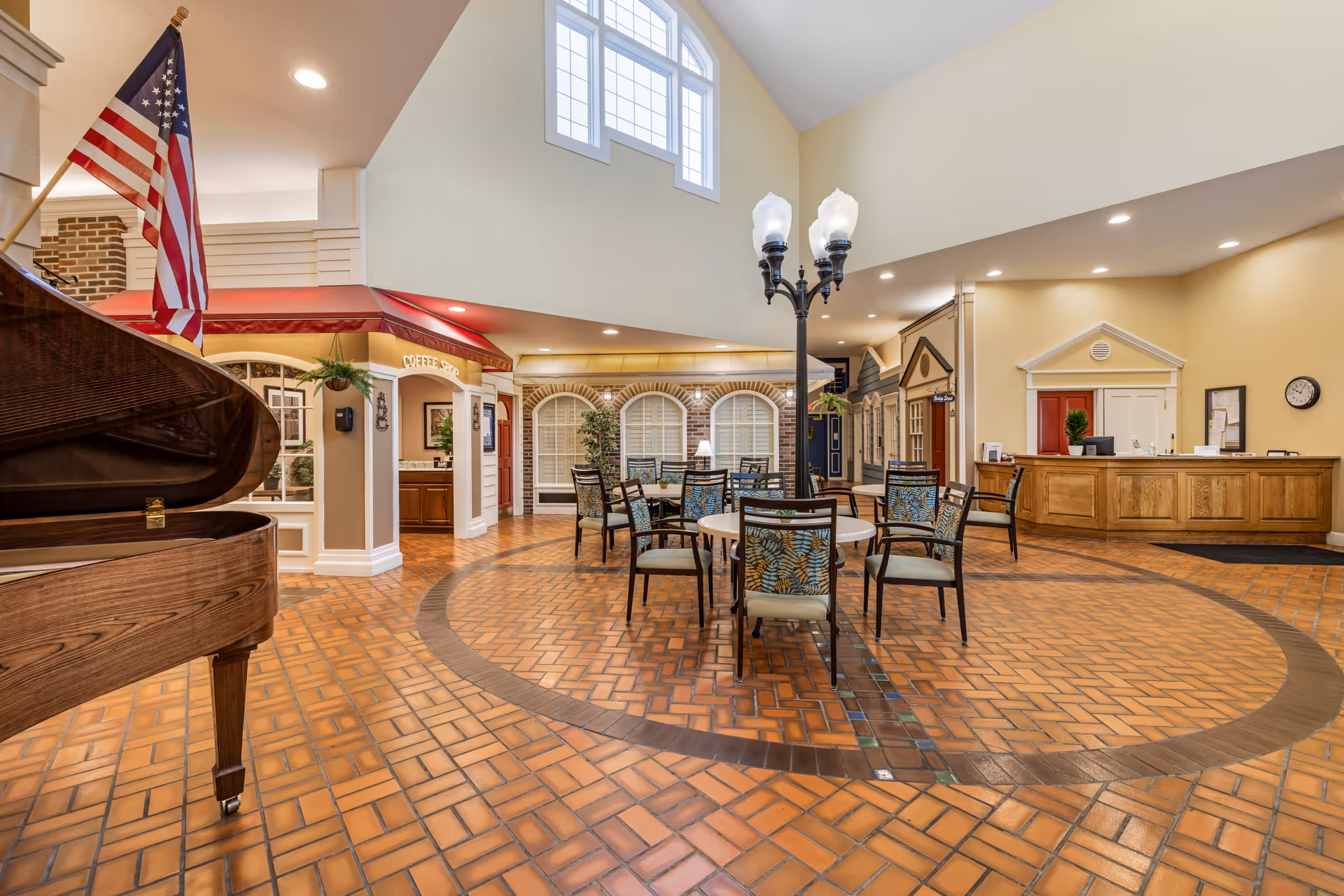 Spacious senior living common area with round tables and chairs, a grand piano, a reception desk and decorative streetlamp under a high skylight.