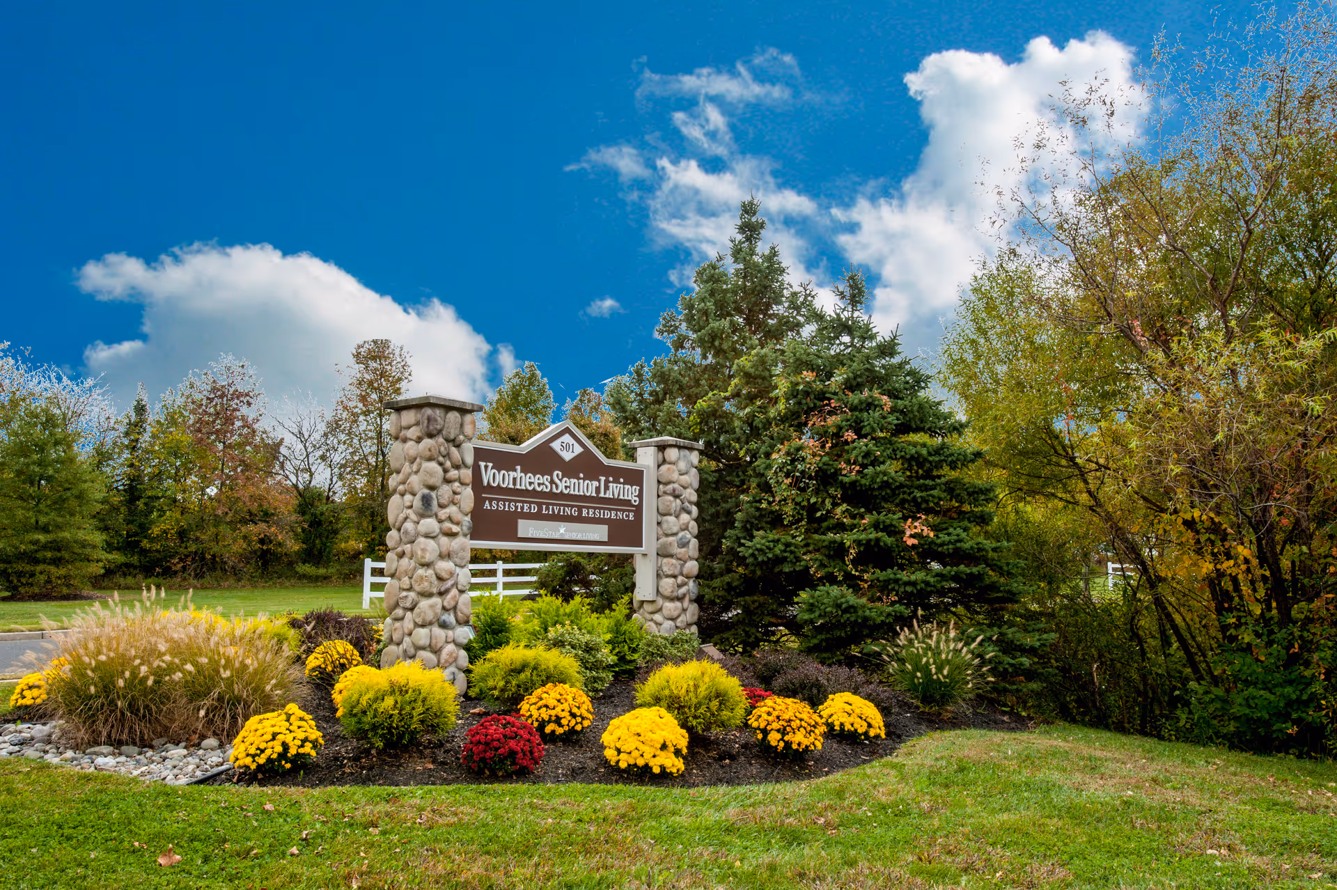 Stone pillars supporting a sign for Voorhees Senior Living Assisted Living Residence surrounded by landscaped bushes, colorful flowers, and trees under a blue sky with scattered clouds.