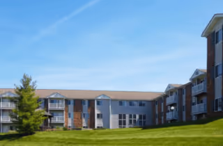 Exterior view of a multi-story senior living facility named Heritage Place with brick and siding facade, multiple windows, balconies, and a well-maintained grassy lawn under a clear blue sky.