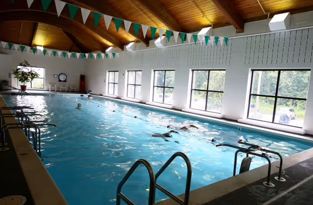 Indoor swimming pool with several swimmers, large windows along one side, a wooden vaulted ceiling and green-and-white lane flags.