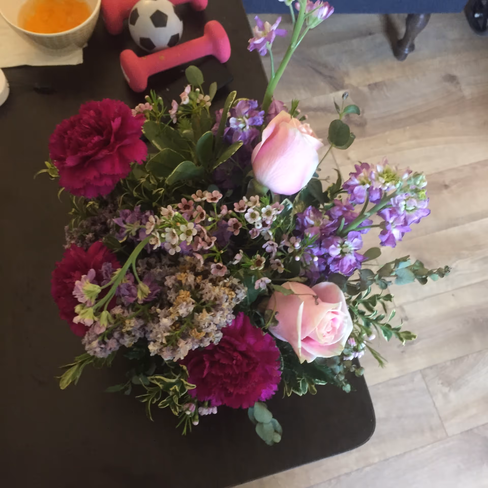 A close-up view of a floral arrangement featuring pink roses, dark pink carnations, and various small purple and white flowers on a dark table. In the background, there are pink dumbbells, a small soccer ball, a bowl with orange liquid, and a wooden floor.