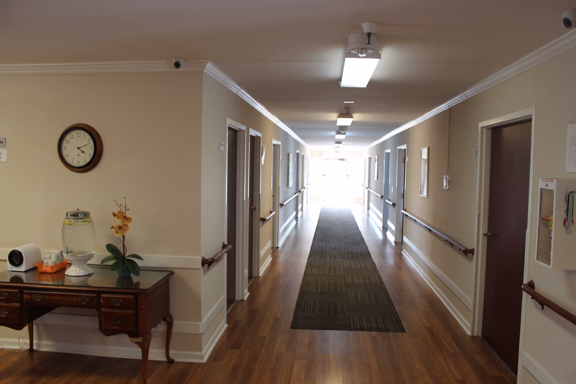 A long, well-lit hallway in a senior living facility with wooden floors, beige walls, handrails on both sides, and several closed doors leading to rooms. A wooden table with a water dispenser, tissue box, and a small plant is positioned on the left side near the entrance of the hallway. A clock is mounted on the wall above the table.
