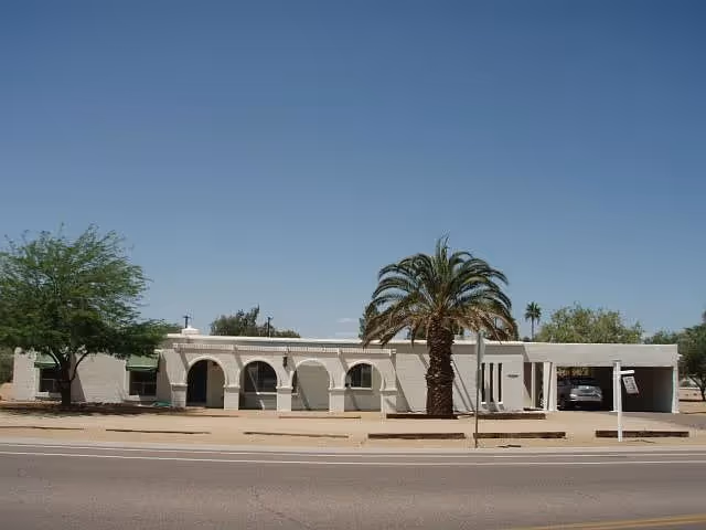 Single-story white stucco building with arched windows and a palm tree in front along a street.