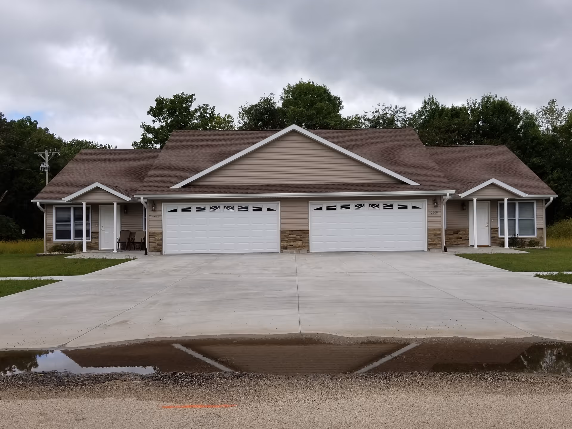 Front exterior view of a duplex building with beige siding, brown roof, two white garage doors, and two front entrances with small porches. There is a concrete driveway and a small puddle reflecting part of the building. Trees and cloudy sky are in the background.