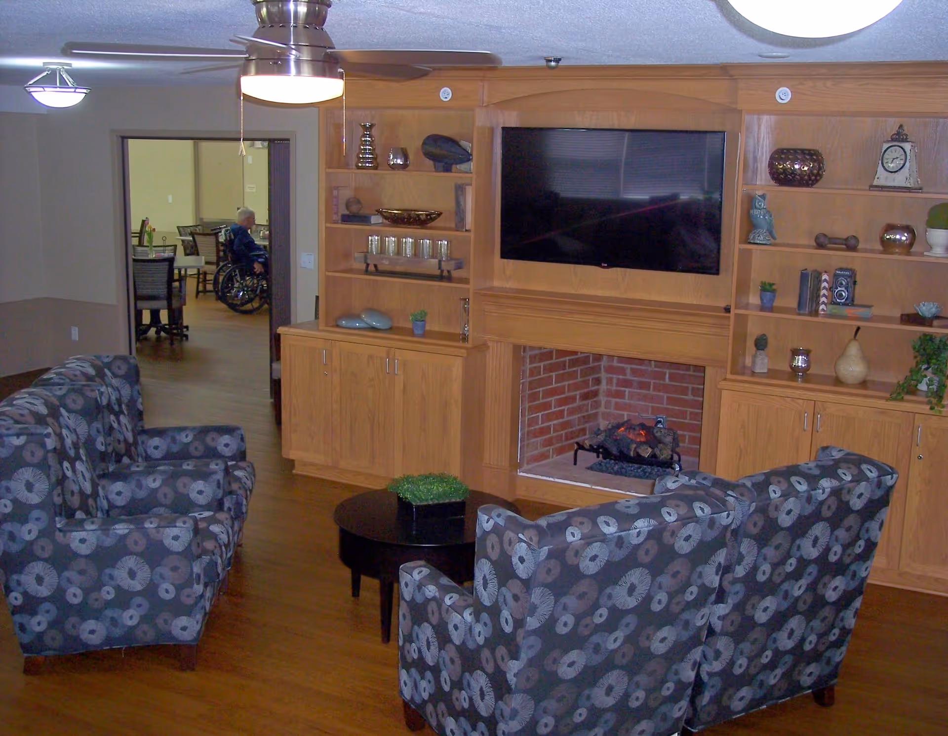 A cozy living room area with patterned armchairs arranged around a small round black table with a green plant. The room features a wooden built-in entertainment center with a flat-screen TV mounted above a brick fireplace. Shelves on either side of the TV hold decorative items including vases, books, and small plants. A ceiling fan with a light fixture hangs above. In the background, a person in a wheelchair is visible in an adjacent room with tables and chairs.