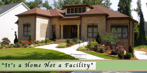 A single-story brick building with a brown shingled roof surrounded by a well-maintained lawn and landscaped garden beds. A concrete walkway leads to the entrance, which has a covered porch with steps and a ramp. Trees and shrubs are visible around the building.