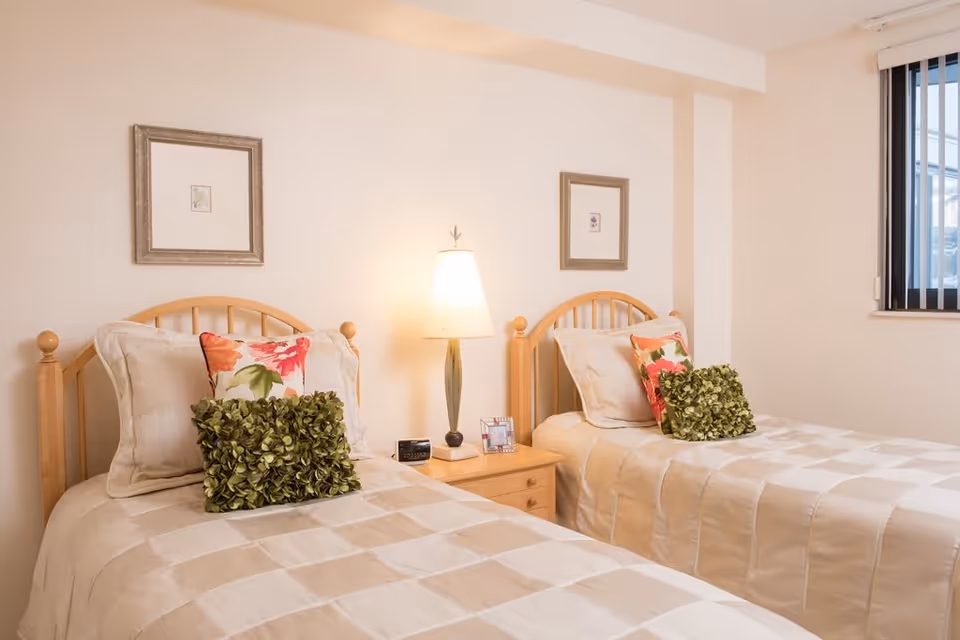 A bedroom with two single beds featuring light wood headboards, beige checkered bedding, and decorative pillows in green and floral patterns. A wooden nightstand with a lamp, clock, and picture frame is placed between the beds. Two framed pictures hang on the wall above each bed. A window with vertical blinds is visible on the right side.