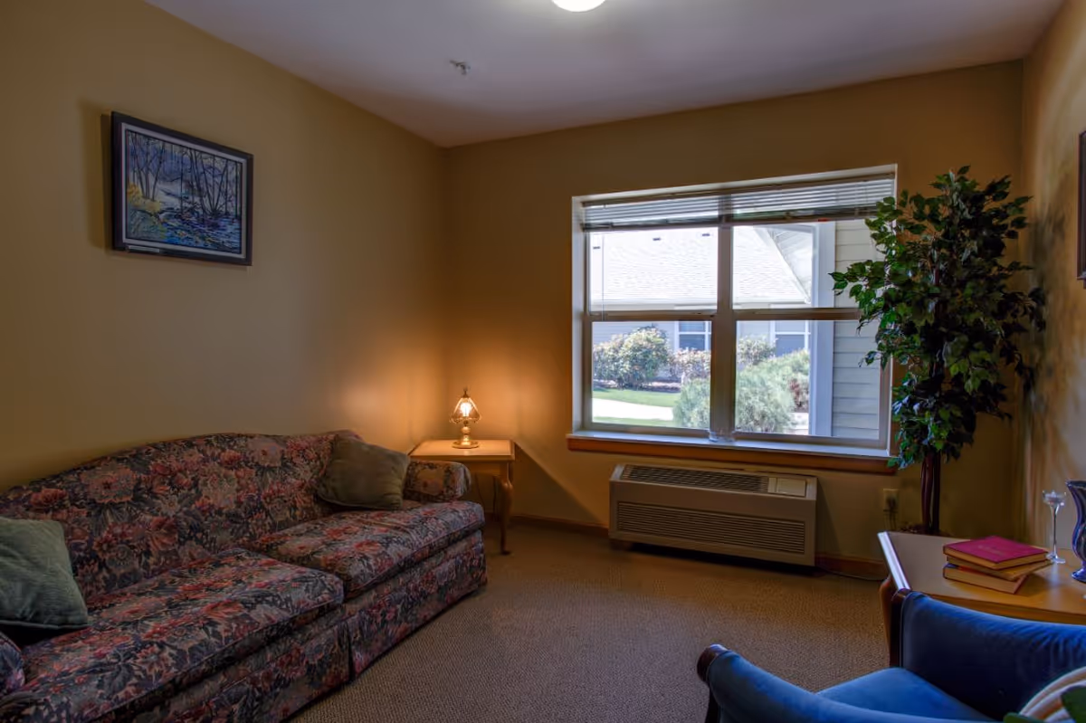 A cozy living room with a floral patterned sofa, a small wooden side table with a lamp, a large window showing greenery outside, a blue armchair, a wooden table with books and a vase, and a tall leafy plant in the corner.
