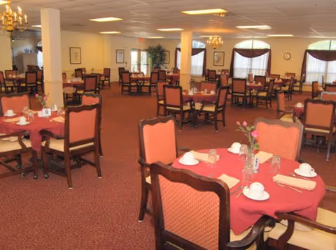 A spacious dining room with multiple tables covered in red tablecloths, each set with white cups, saucers, glasses, and napkins. The chairs have wooden frames with red upholstered backs and seats. The room has carpeted floors, chandeliers, large windows with curtains, and framed pictures on the walls.