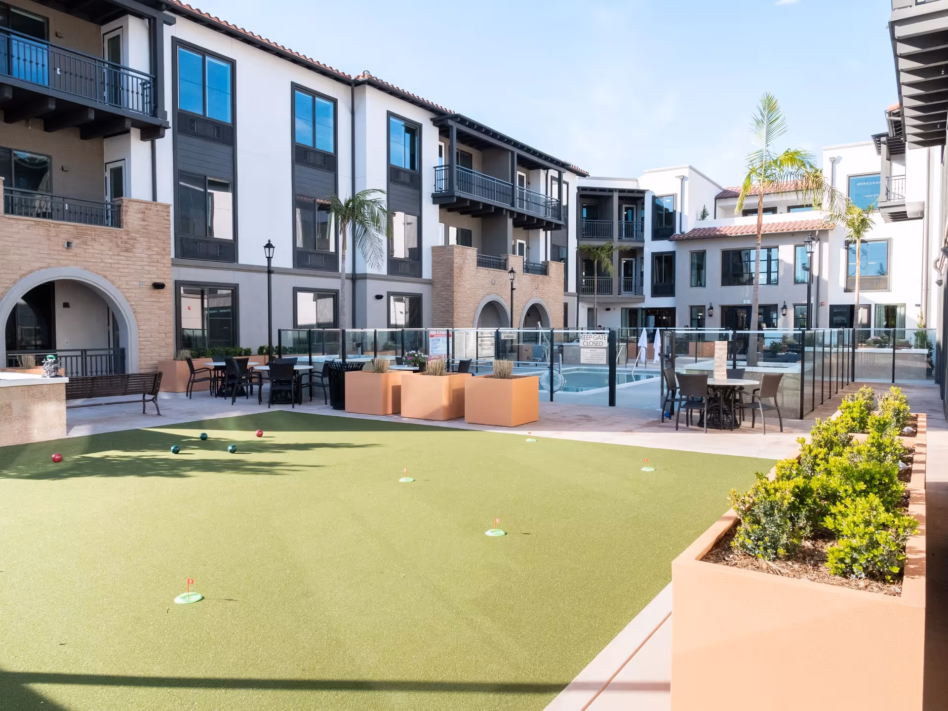 A bright courtyard with an artificial putting green, outdoor tables and chairs, a fenced pool, and surrounding three-story Mediterranean-style buildings.