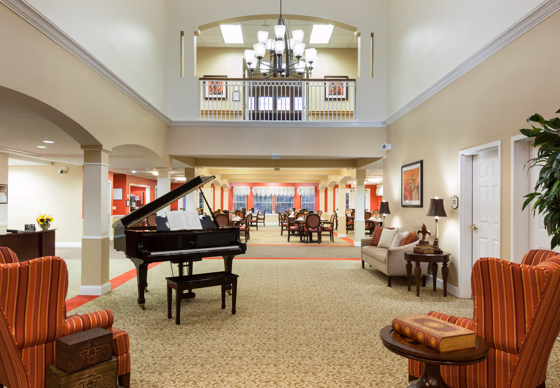 Spacious and well-lit senior living facility common area featuring a grand piano with sheet music, comfortable seating including striped armchairs and a beige sofa, decorative lamps, framed artwork on the walls, and a dining area with multiple tables and chairs in the background. The space has high ceilings with a chandelier and a balcony overlooking the area.