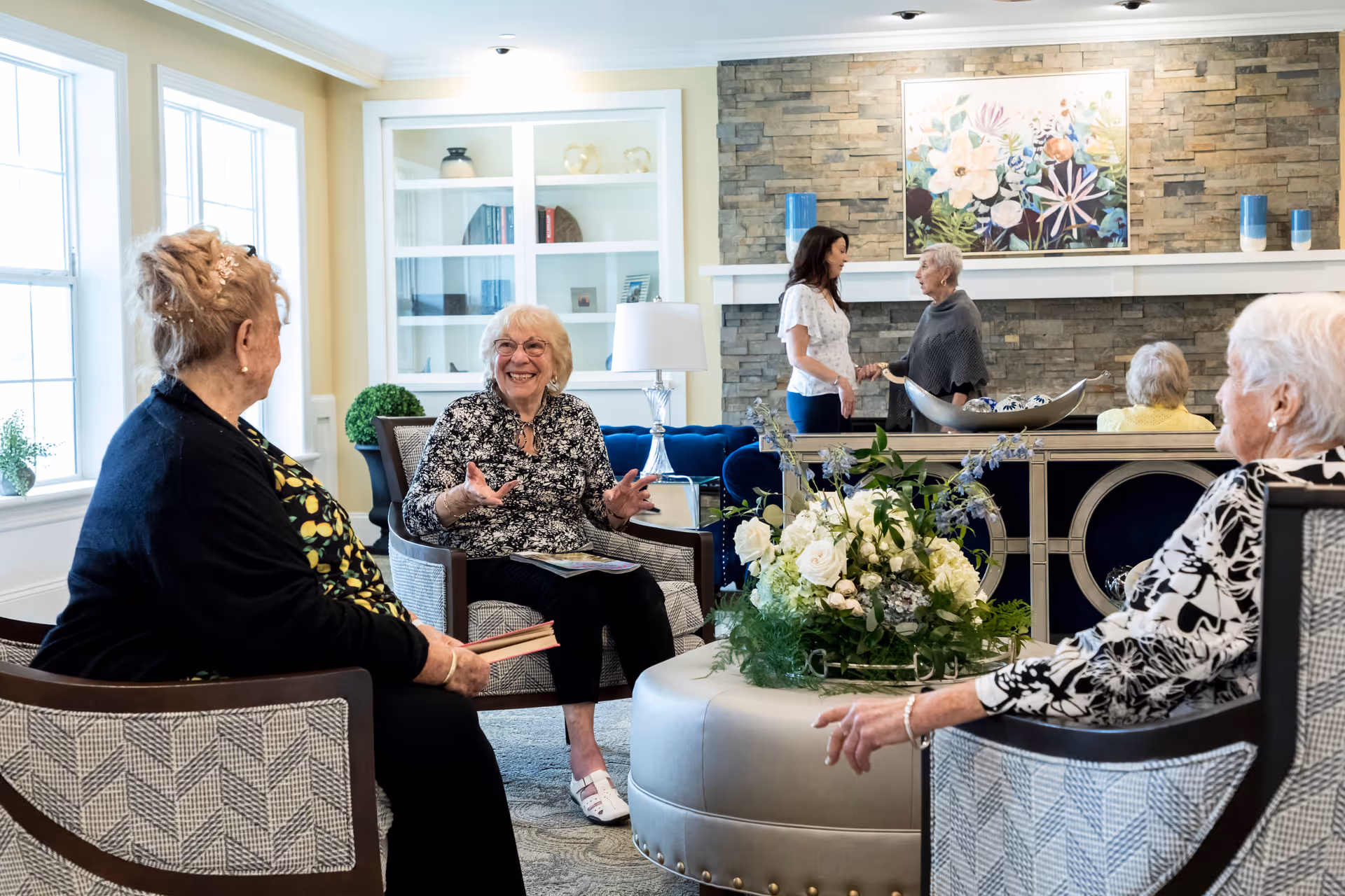 A cozy living room in the Villas of Holmdel facility with four elderly women sitting and chatting around a round ottoman decorated with a floral arrangement. In the background, two women are standing and conversing near a stone fireplace with a colorful floral painting above it. The room has large windows, a built-in white cabinet, and comfortable patterned chairs.