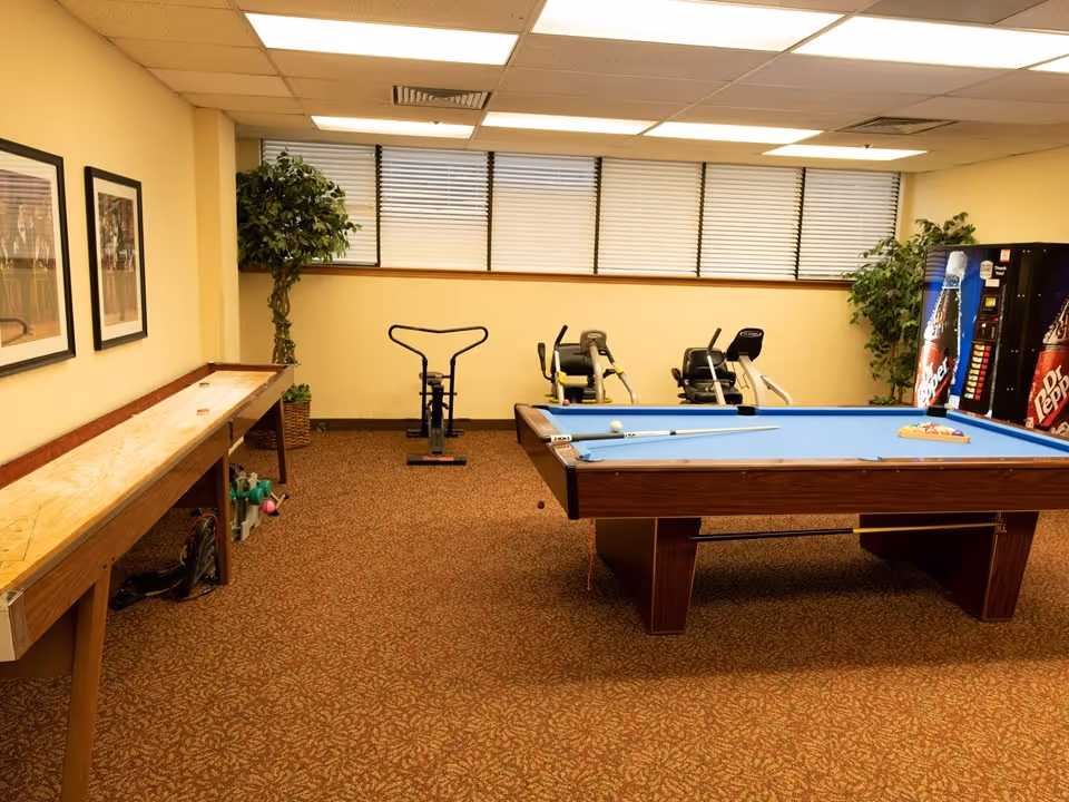 A recreational room with a blue pool table in the center, a shuffleboard table on the left, two exercise bikes against the back wall, and two vending machines on the right. The room has beige walls, a brown patterned carpet, framed pictures on the left wall, and two artificial plants near the back wall windows with closed blinds.