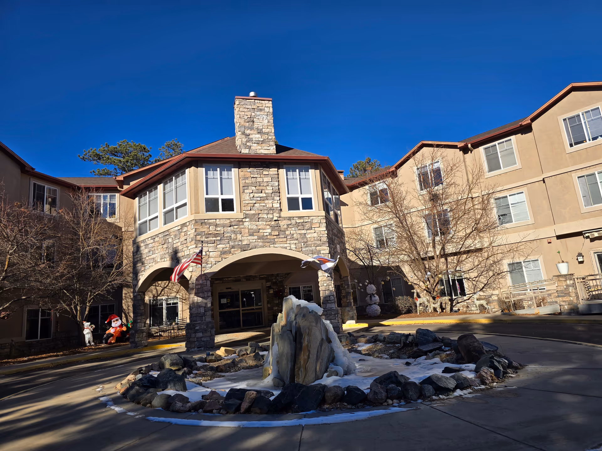 Exterior view of Elk Run Assisted Living facility showing a stone and beige building with multiple windows, a central entrance with an archway, two flags, and a circular driveway with a rock and ice fountain in the middle. There are leafless trees and holiday decorations including a Santa figure and a snowman near the entrance.