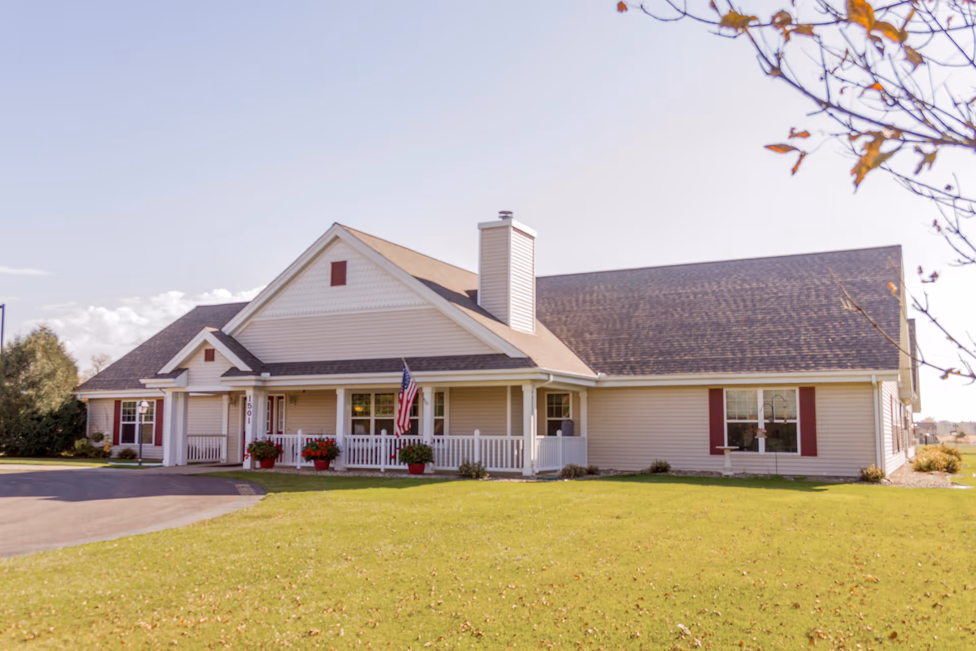 Exterior view of a single-story residential building with beige siding, a dark shingled roof, and a white front porch railing. There is an American flag hanging on the porch, several potted plants with red flowers, and a well-maintained green lawn in front. Trees with some autumn leaves frame the image.