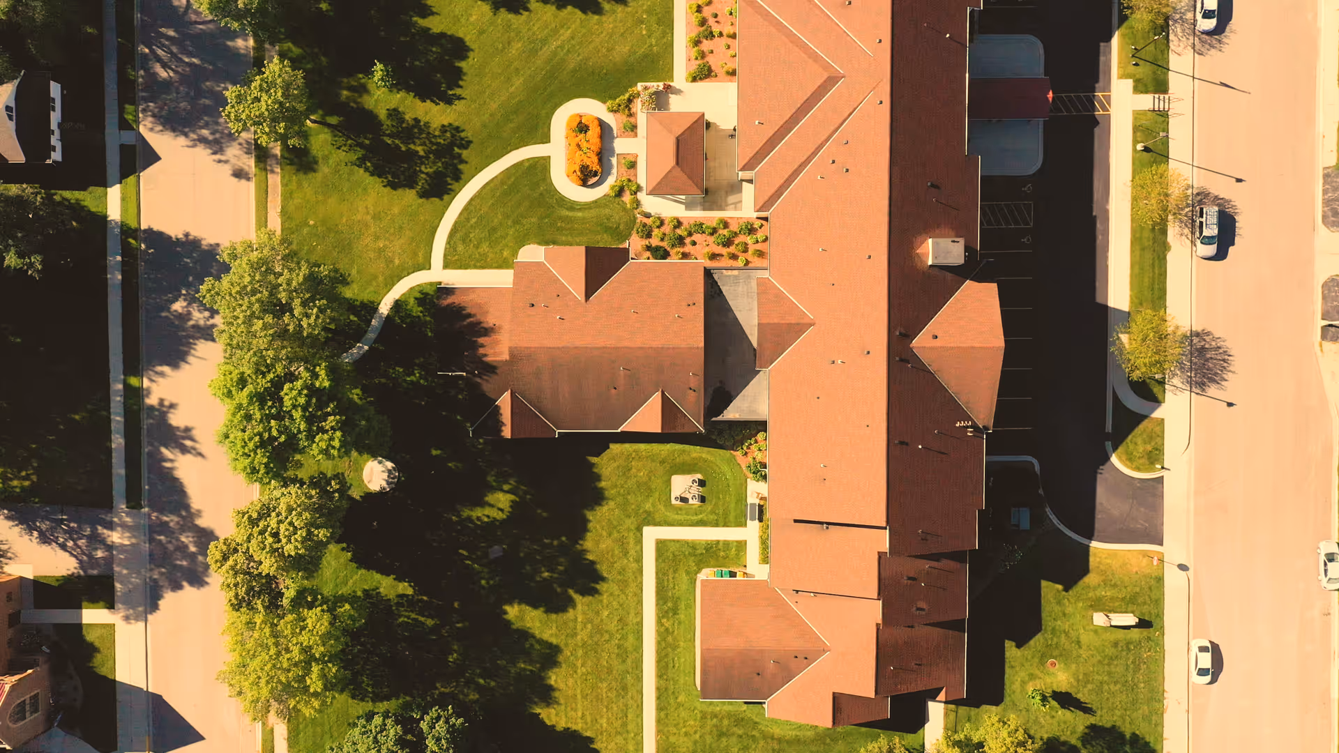 Aerial view of a long red-roofed senior living building with surrounding lawns, walking paths, trees and parked cars.