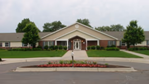 Front view of a single-story memory care facility with a central entrance, landscaped lawn, and circular driveway.