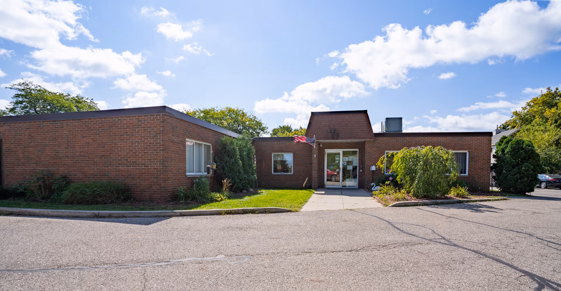 Exterior view of a single-story brick building with a flat roof under a partly cloudy blue sky. The building has a main entrance with glass doors, an American flag mounted near the entrance, and surrounding greenery including bushes and trees. There is a paved driveway or parking area in front of the building.