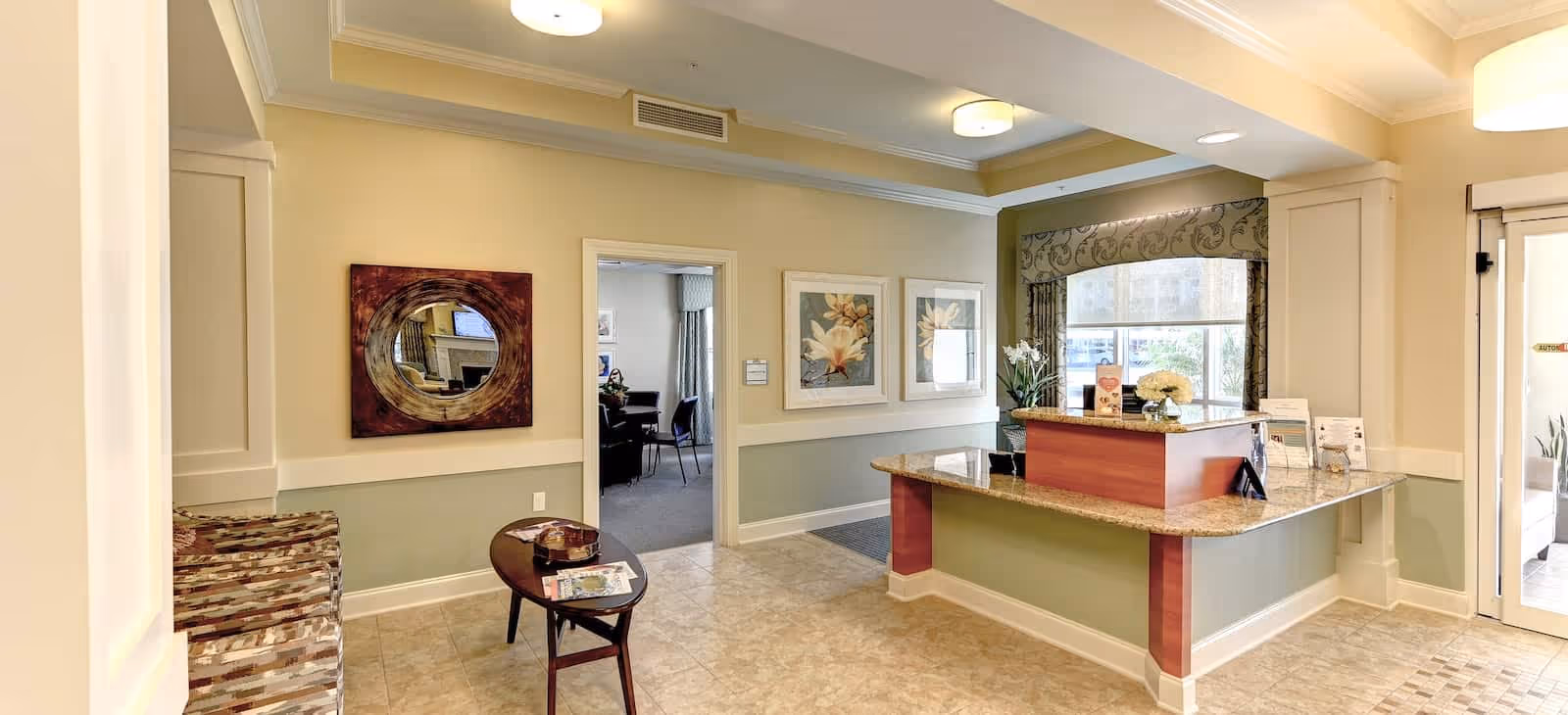 Reception area of a senior living facility with a granite countertop desk, decorative flowers, and informational brochures. The walls are painted in soft beige and green tones, adorned with framed floral artwork and a round mirror. There is a small wooden table with magazines and a patterned bench near the entrance.