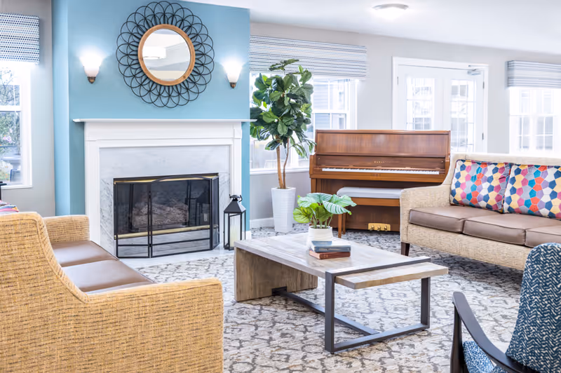 Bright common living room with wicker sofas, a coffee table, a fireplace with decorative mirror, and an upright piano.