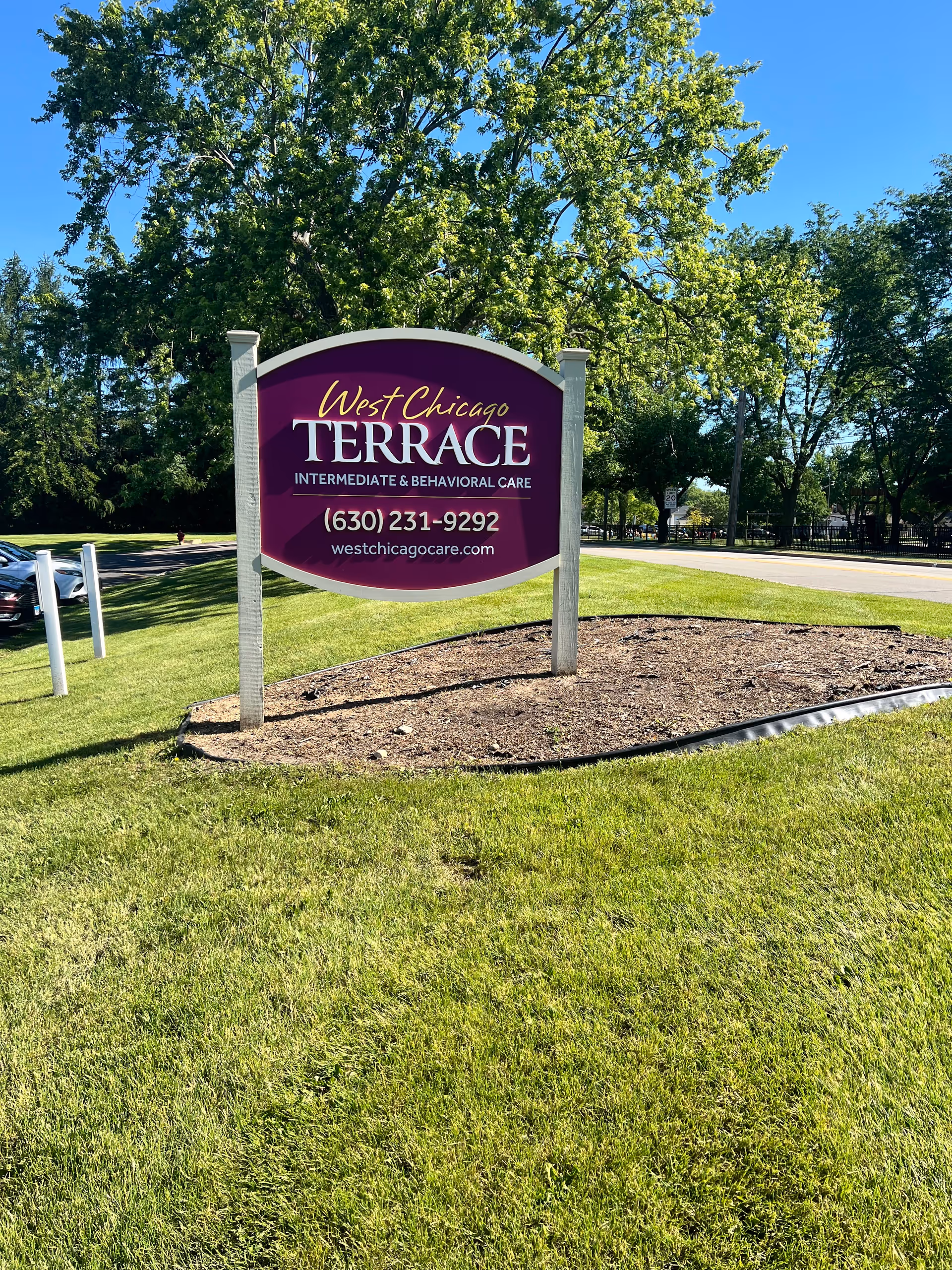 A purple and white sign for West Chicago Terrace Intermediate & Behavioral Care standing on a grassy area with trees and a road in the background under a clear blue sky.