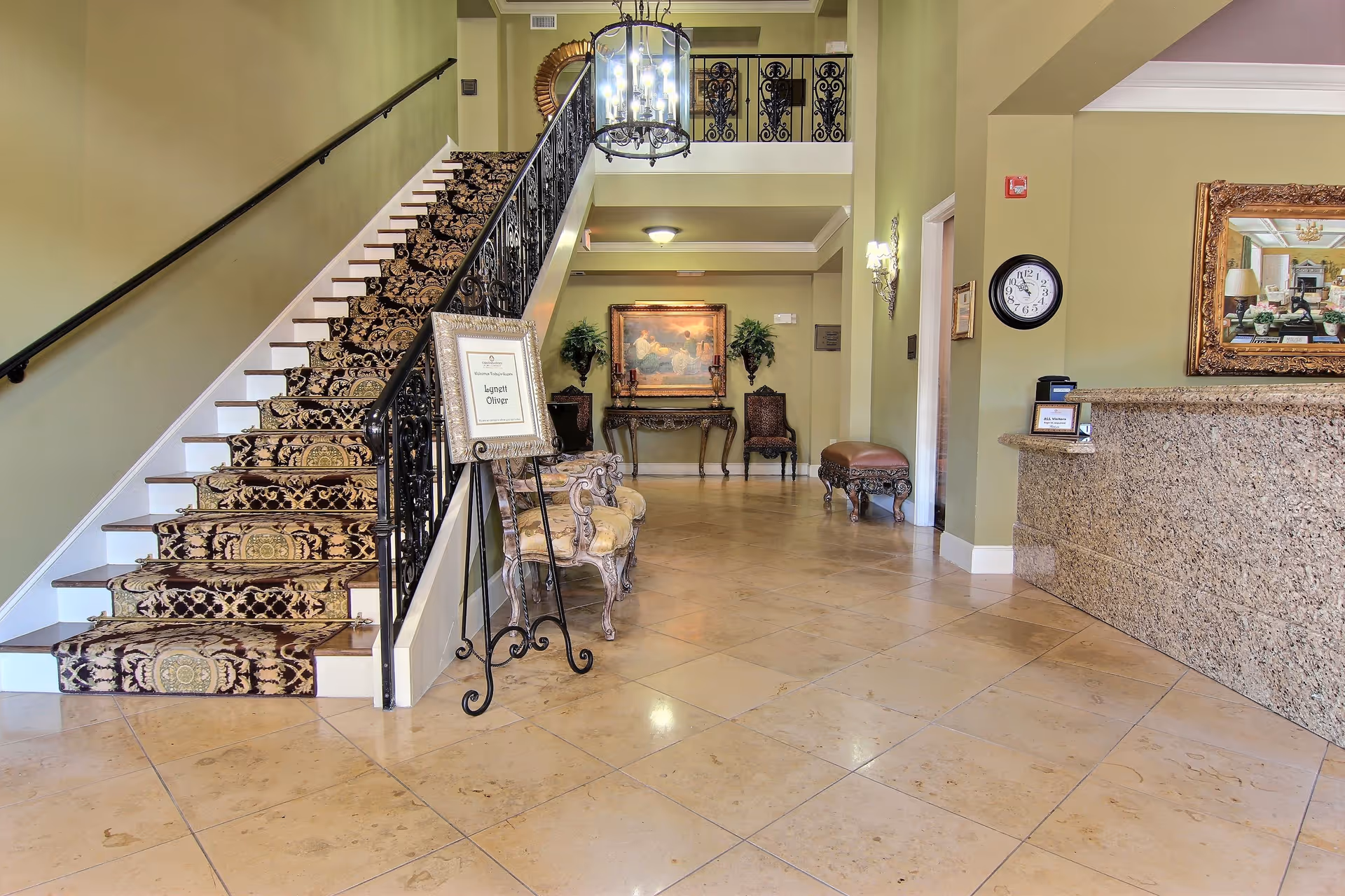 Interior view of a senior living facility lobby with a staircase on the left covered in a patterned carpet runner, a chandelier hanging from the ceiling, a granite reception desk on the right, and decorative chairs and artwork along the back wall.
