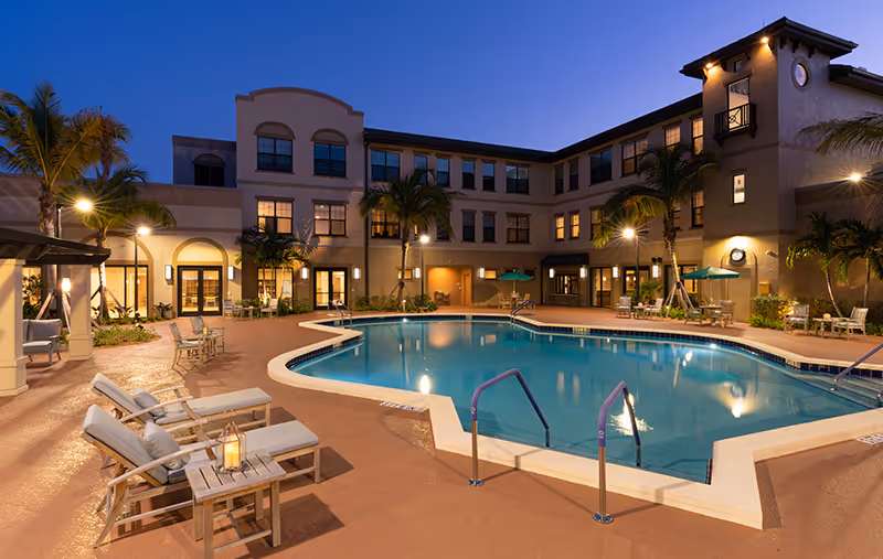 Outdoor swimming pool area at dusk with lounge chairs, tables, and umbrellas surrounding the pool. The multi-story Abbey Delray Health Care Center building is visible in the background with palm trees and outdoor lighting.