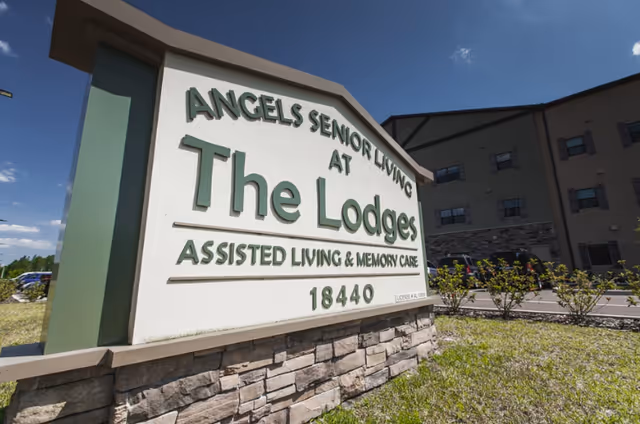 Outdoor view of a large sign for Angels Senior Living at The Lodges, an assisted living and memory care facility, with the building and blue sky in the background.