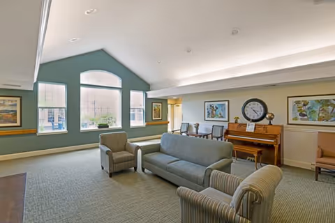 Spacious senior living common room with sofas and armchairs, a piano, large arched windows, and framed artwork.
