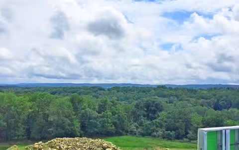 A scenic view of a lush green forest under a partly cloudy blue sky, with a pile of rocks and part of a building structure visible in the foreground.