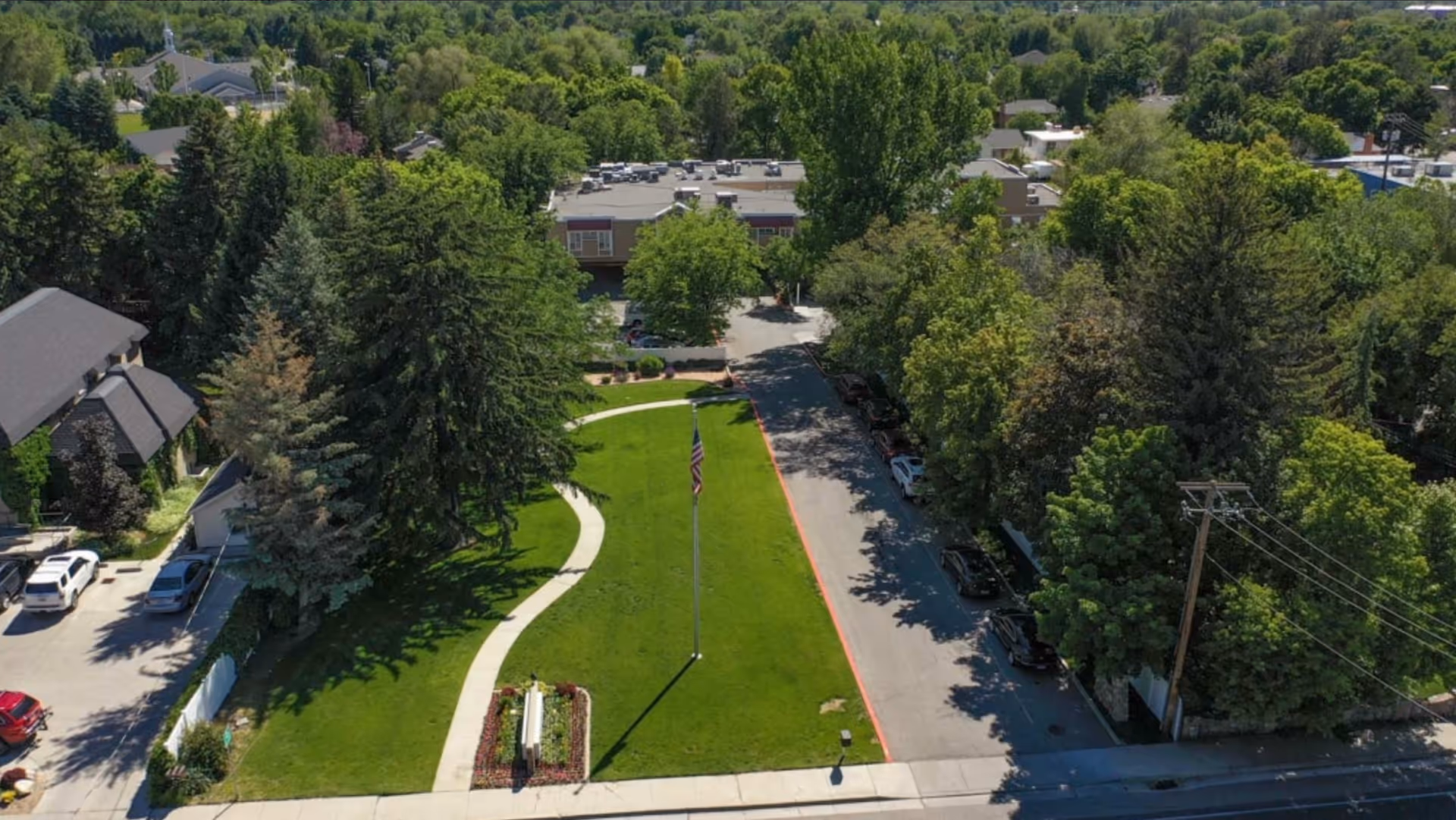 Aerial view of a healthcare facility's outdoor area with a large green lawn, a curved walkway, an American flag on a flagpole, surrounded by trees and buildings, with parked cars along the driveway.