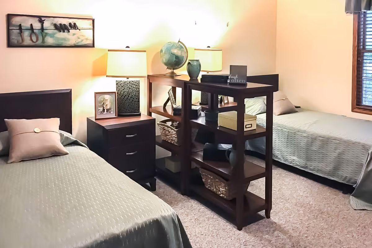 A cozy bedroom with two single beds separated by a dark wooden shelving unit. Each bed has a light green bedspread and a pillow. Between the beds is a nightstand with a lamp, a framed photo, and decorative items. The room has beige walls, a window with wooden blinds, and a wall decoration above one bed.