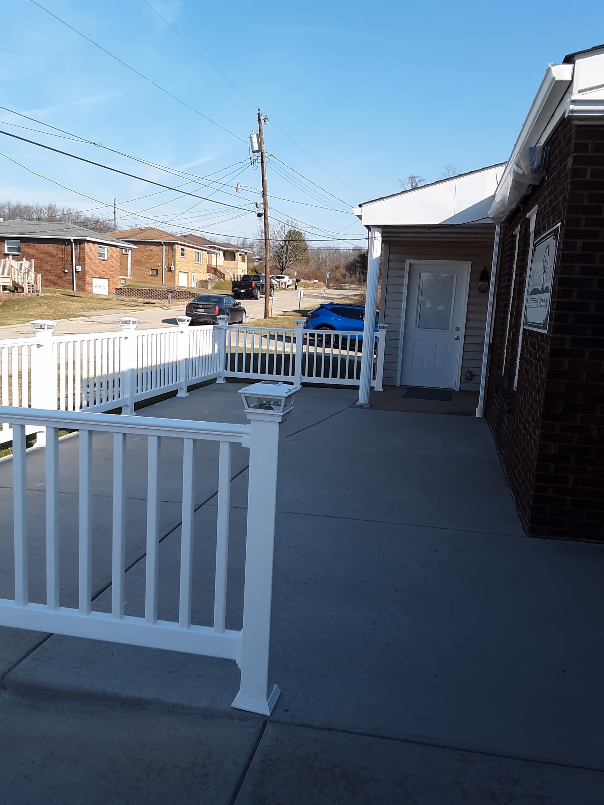 Front entrance and ramp of a brick assisted living building with white railings, a concrete porch, and a street view with parked cars.