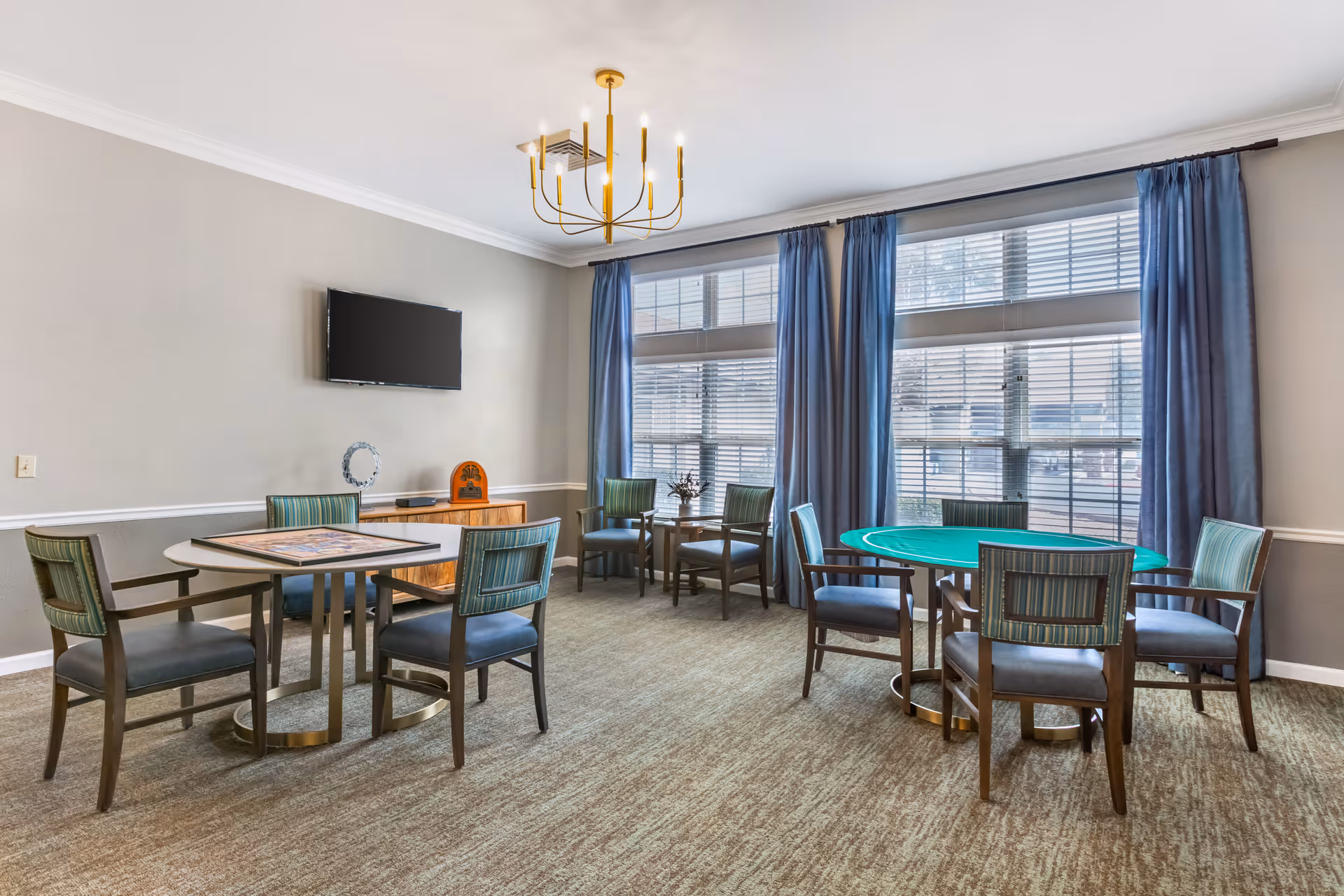 A well-lit common room with two round tables surrounded by chairs. One table has a green felt surface, possibly for card games, and the other has a puzzle on it. Large windows with blue curtains let in natural light. A flat-screen TV is mounted on the wall above a wooden cabinet, and a modern gold chandelier hangs from the ceiling.