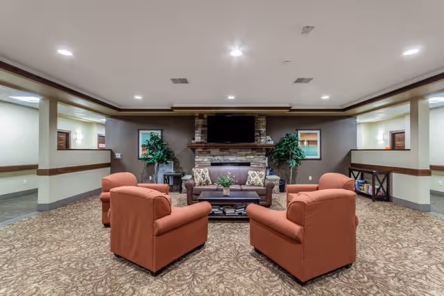 A cozy living room area with four orange armchairs arranged around a brown sofa and a wooden coffee table. Behind the sofa is a stone fireplace with a mounted flat-screen TV above it. Two green potted plants flank the fireplace, and framed artwork hangs on the wall. The room has recessed ceiling lights and patterned carpet flooring.