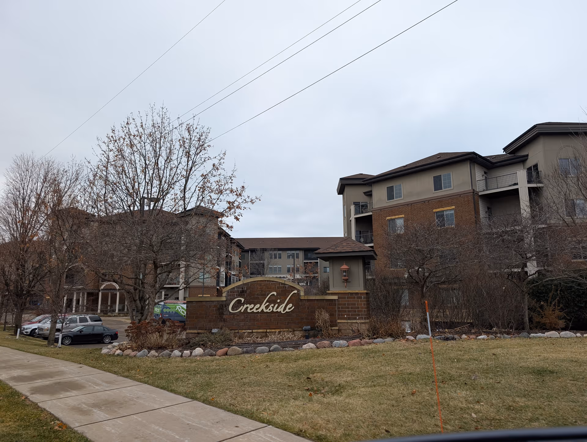 Exterior view of Creekside in Edina senior living facility showing a brick sign with the name 'Creekside' in front of a multi-story building with balconies, leafless trees, parked cars, and a cloudy sky.