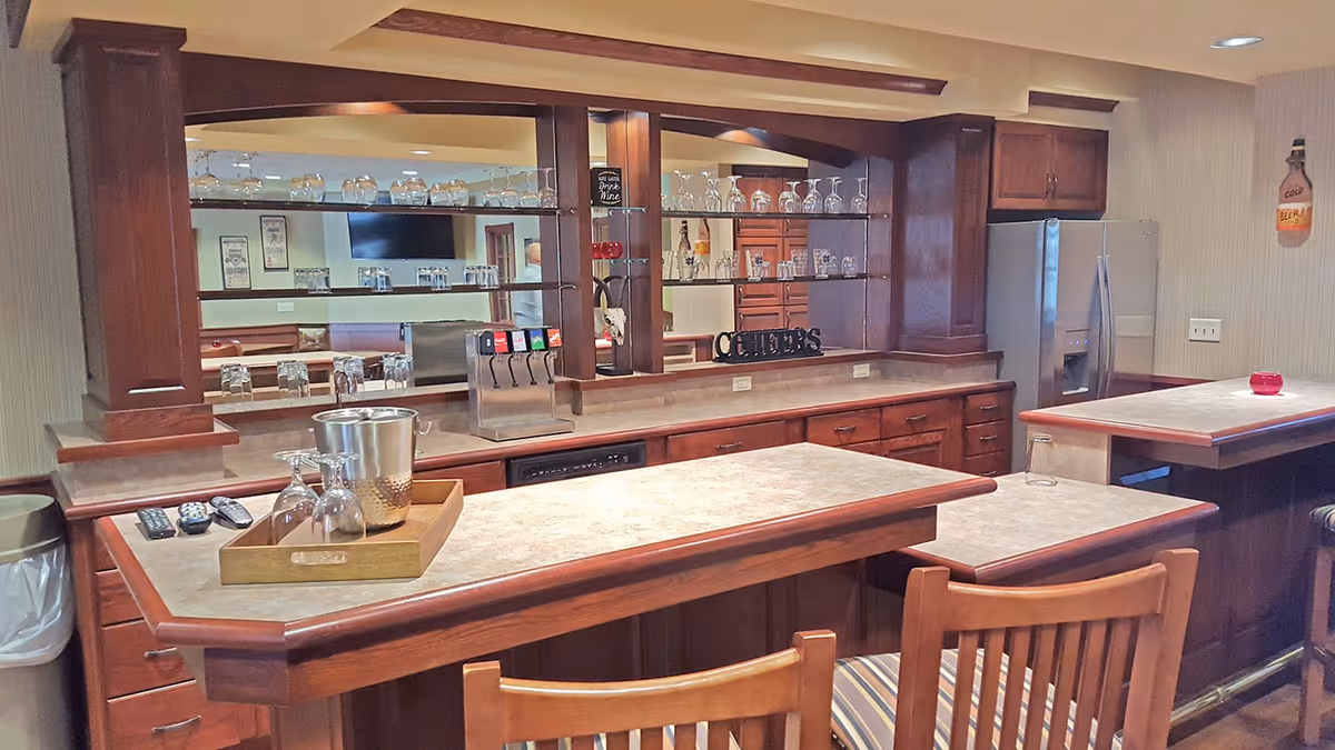 Interior view of a bar area in a retirement community featuring a countertop with a wooden tray holding an ice bucket and glasses, a soda fountain machine, glass shelves with various glasses, wooden cabinetry, a refrigerator, and a wall decoration shaped like a bottle.