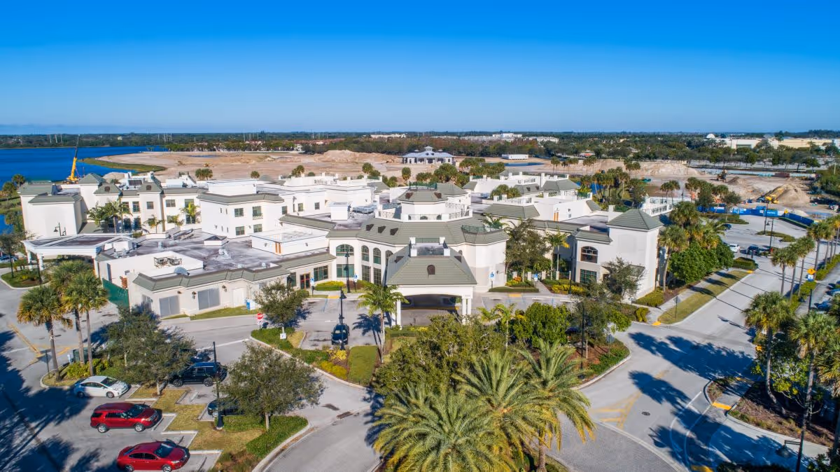 Aerial view of Luxe Senior Living at Wellington facility showing multiple white buildings with green roofs surrounded by palm trees and landscaped greenery. The facility is located near a body of water and is bordered by roads with parked cars and construction visible in the background under a clear blue sky.