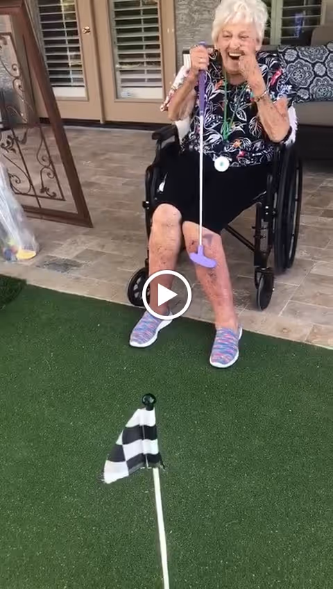 An elderly woman sitting in a wheelchair on a patio, holding a purple mini golf club and laughing. In front of her is a putting green with a black and white checkered flag marking the hole. The patio has tiled flooring and a cushioned bench with patterned pillows in the background.
