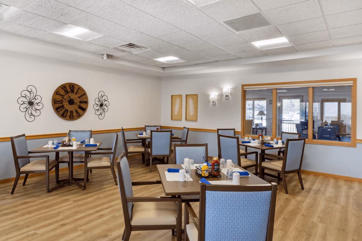 A dining room in a senior living facility with several square tables set with white cups, napkins, and condiments. The room has light wood flooring, light blue and beige chairs, and neutral-colored walls decorated with a large wooden clock and wall art. A large window looks into another room with additional seating.
