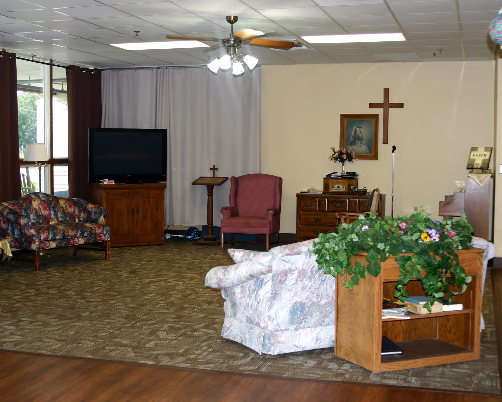 A senior living facility lounge with upholstered sofas and armchairs, a TV and wooden cabinets, a floral planter, and a cross on the wall.