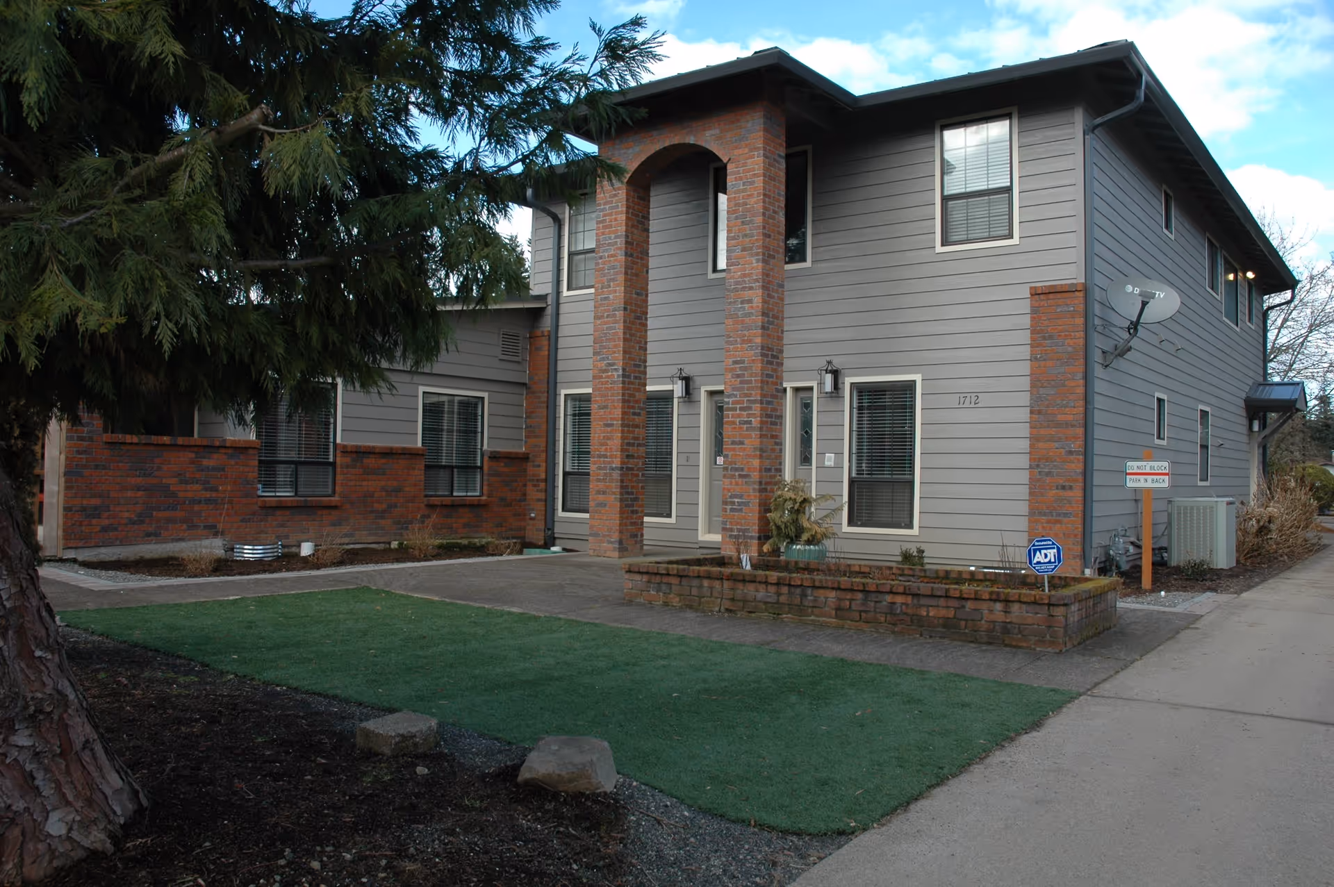 Two-story gray siding and brick building with an arched double-column entrance, small front yard and an ADT security sign.