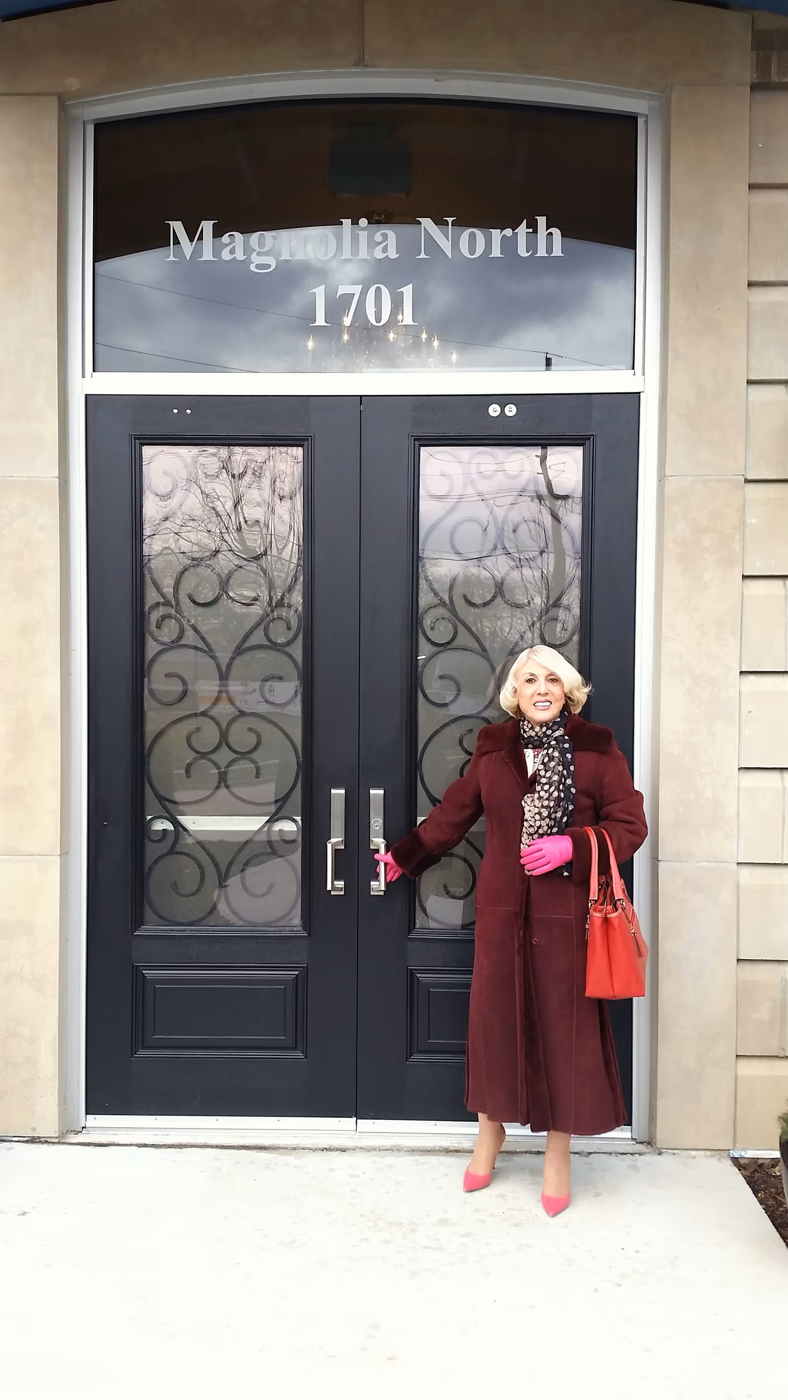 A woman in a burgundy coat and pink gloves stands at the entrance of Magnolia North, smiling as she holds the door handle. The entrance features elegant black doors with decorative glass panels and the name 'Magnolia North' displayed above.