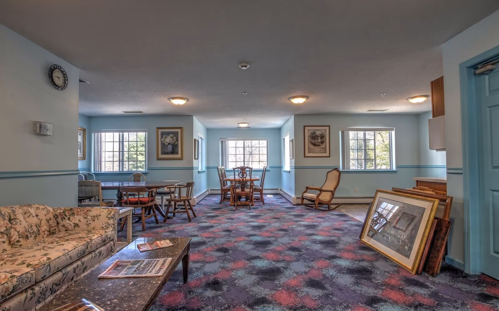 A spacious common area in a senior living facility with light blue walls and a patterned carpet in shades of purple, pink, and blue. The room features a floral upholstered sofa, a marble-top coffee table with magazines, several wooden dining tables and chairs, framed artwork on the walls, and large windows letting in natural light. There are also some framed pictures leaning against the wall near a blue door.