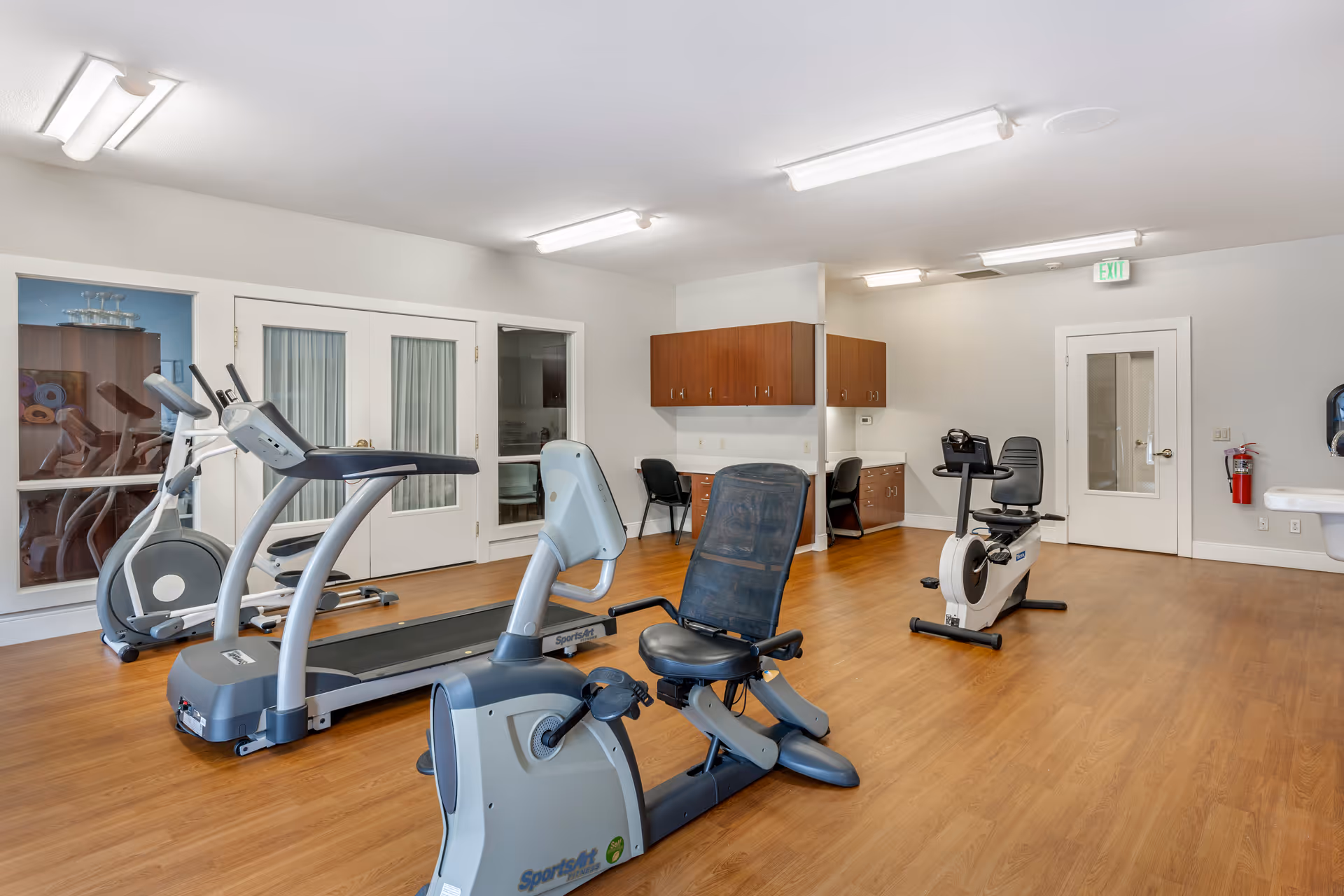 A fitness room with exercise equipment including a treadmill, two recumbent stationary bikes, and an elliptical machine. The room has wood flooring, white walls, overhead fluorescent lighting, and built-in wooden cabinets with two chairs and desks underneath. There is a door with glass panels and an exit sign above another door in the background.