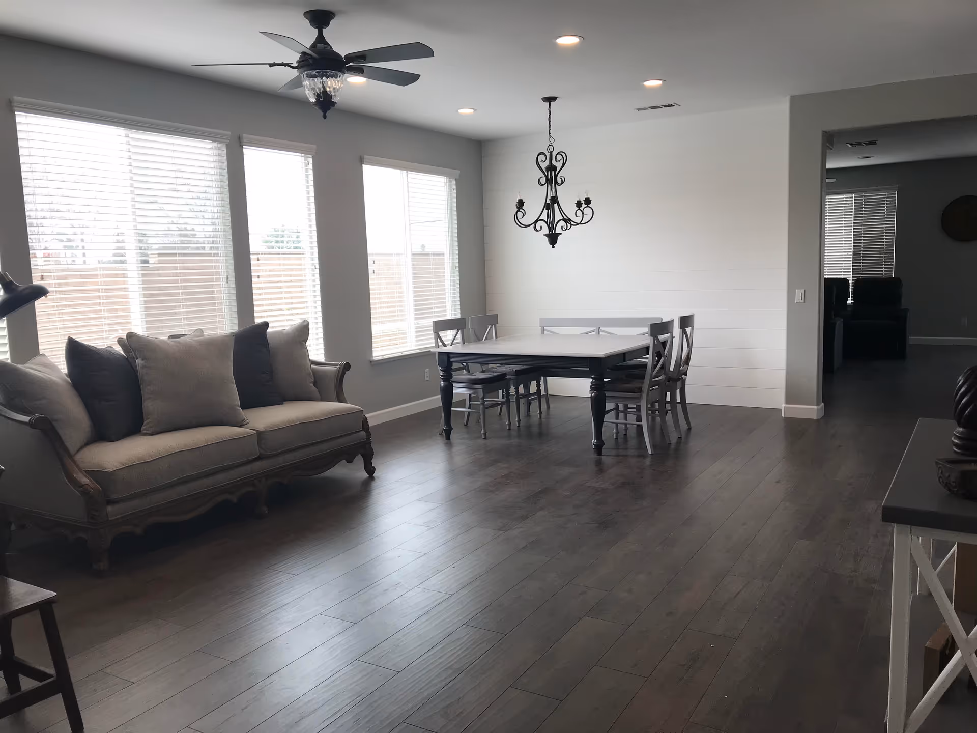 A bright interior room with large windows covered by white blinds. The room features a vintage-style sofa with multiple cushions on the left and a dining table with six chairs on the right. A black ceiling fan with lights is mounted on the ceiling, along with recessed lighting and a decorative chandelier above the dining table. The floor is made of dark wood, and the walls are painted light gray.