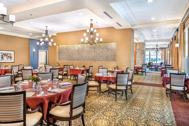 A bright dining room with round tables dressed in red tablecloths, striped-back chairs, chandeliers, floral centerpieces, and patterned carpet.