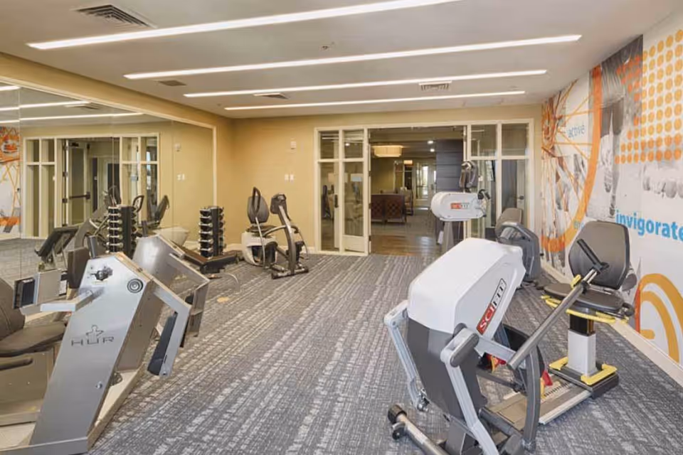 A senior living facility fitness room with exercise machines, including stationary bikes and strength training equipment, a mirrored wall on the left, and a colorful mural on the right wall. The room has carpeted flooring and recessed ceiling lights, with a glass door leading to another area.