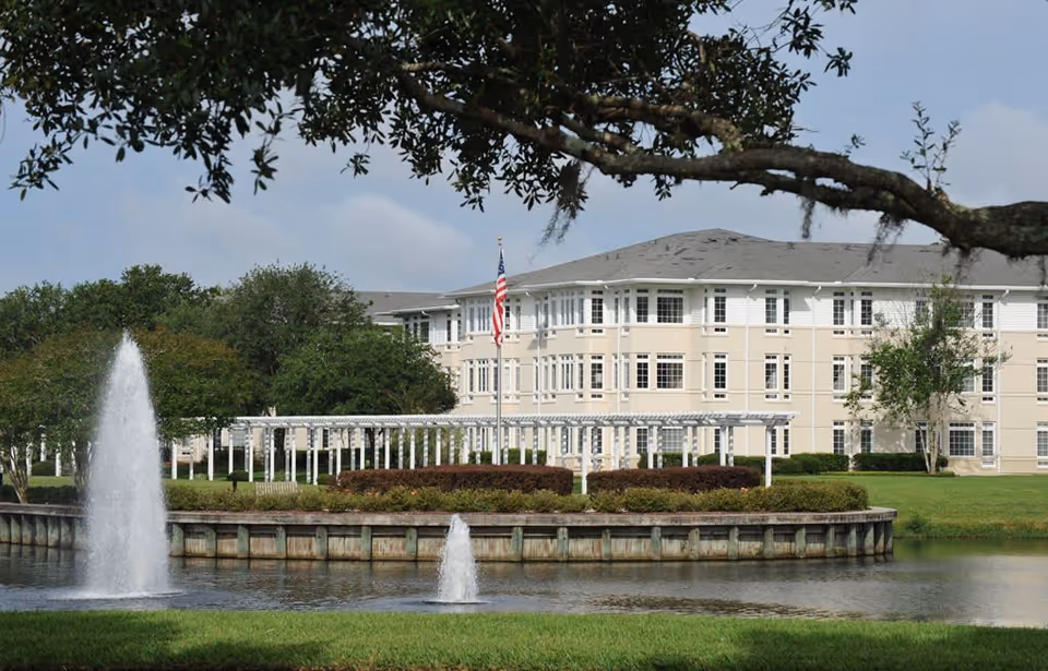 View of a large beige and white multi-story building behind a pond with two water fountains. There is a white pergola structure and an American flag on a flagpole in front of the building. Trees and green grass surround the pond and building, with a large tree branch extending across the top of the image.